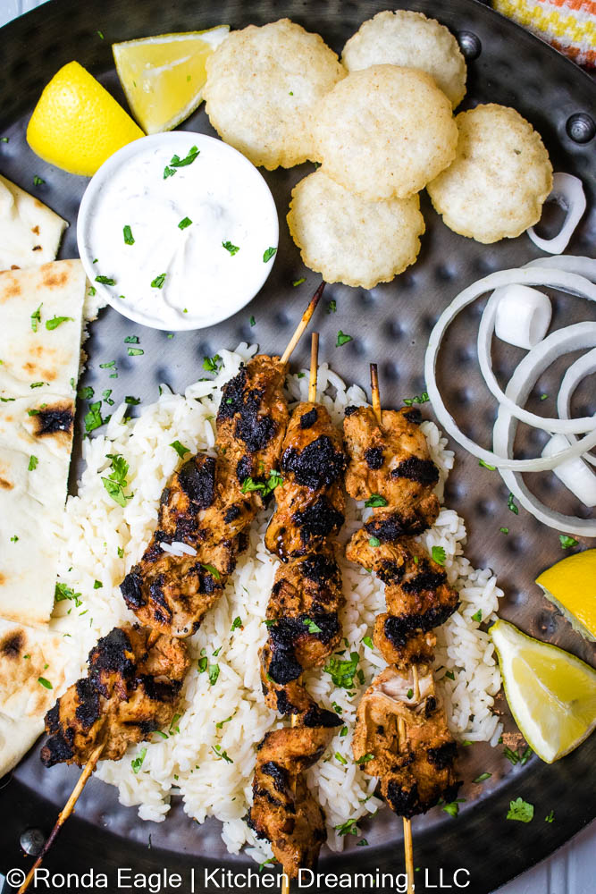  A black hammered metal serving tray filled with Grilled CHicken skewers on a bed of rice, sweet onions, lemon wedges, and raita.