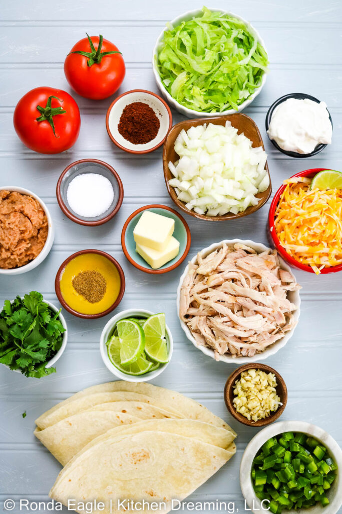 The ingredients for baked chimichangas: tomatoes, lettuce,sour cream, shredded cheese, shredded chicken, diced onions, retired beans, butter, herbs and spices. For a full list, visit the recipe card.