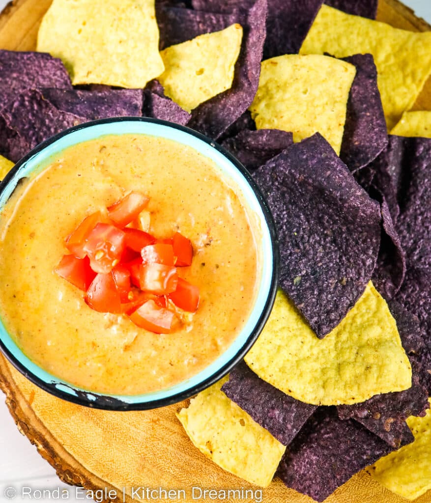 A bowl of nacho dip surrounded by purple and yellow corn chips.
