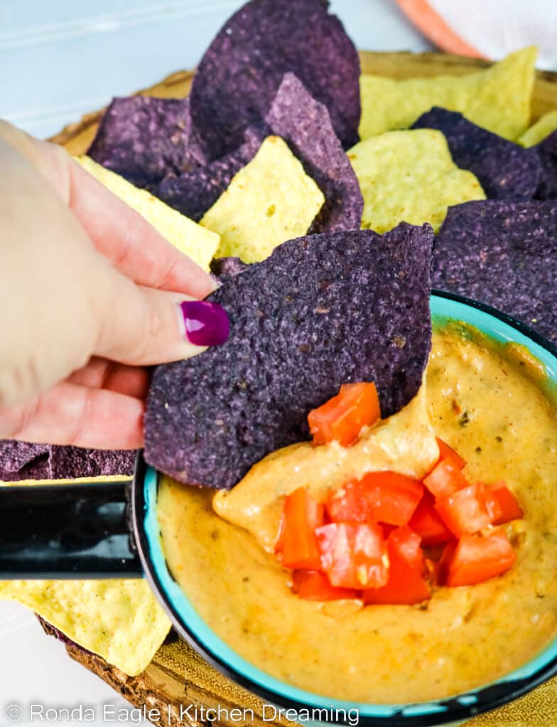 A bowl of nacho cheese dip with purple and yellow corn chips. A hand is dipping a chip into the bowl. No double dipping!