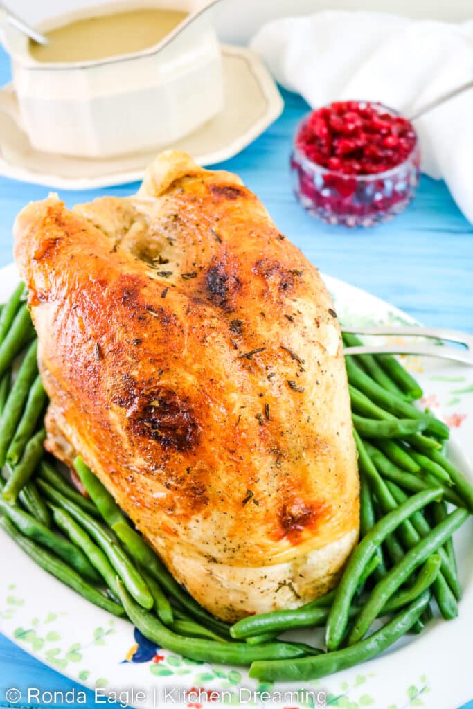 A slow-cooked turkey breast on a platter surrounded by green beans. In the background, there is a gravy boat filled with pan gravy and a dish of cranberry relish.