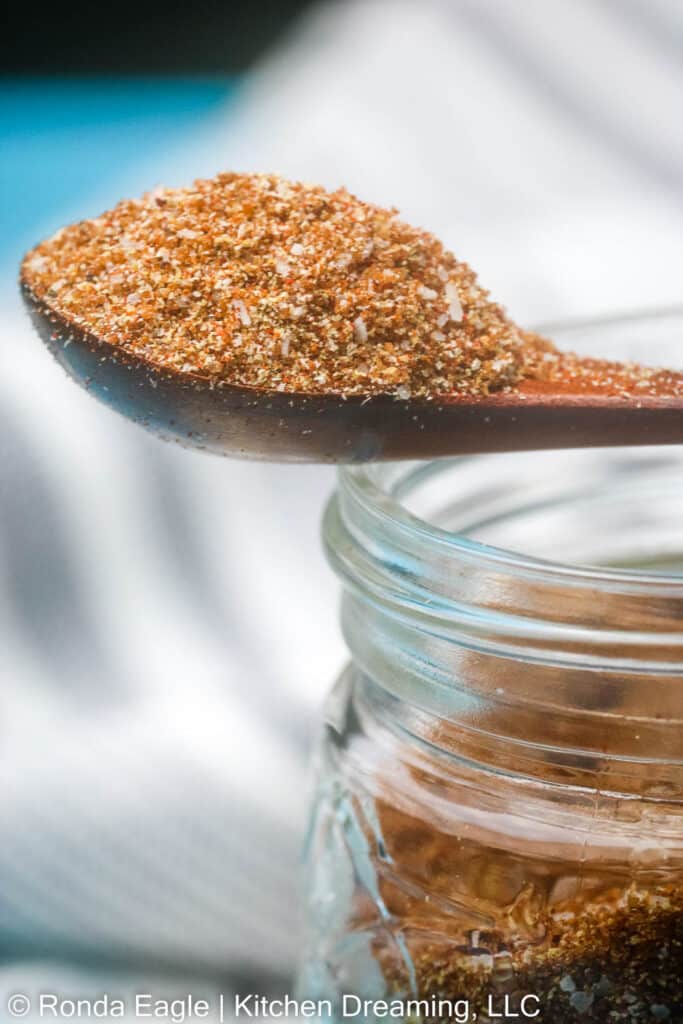 A close up image of the side shot of a wooden spoon filled with a heaping amount of Sweet BBQ rub which is resting across the top of a small glass storage jar.