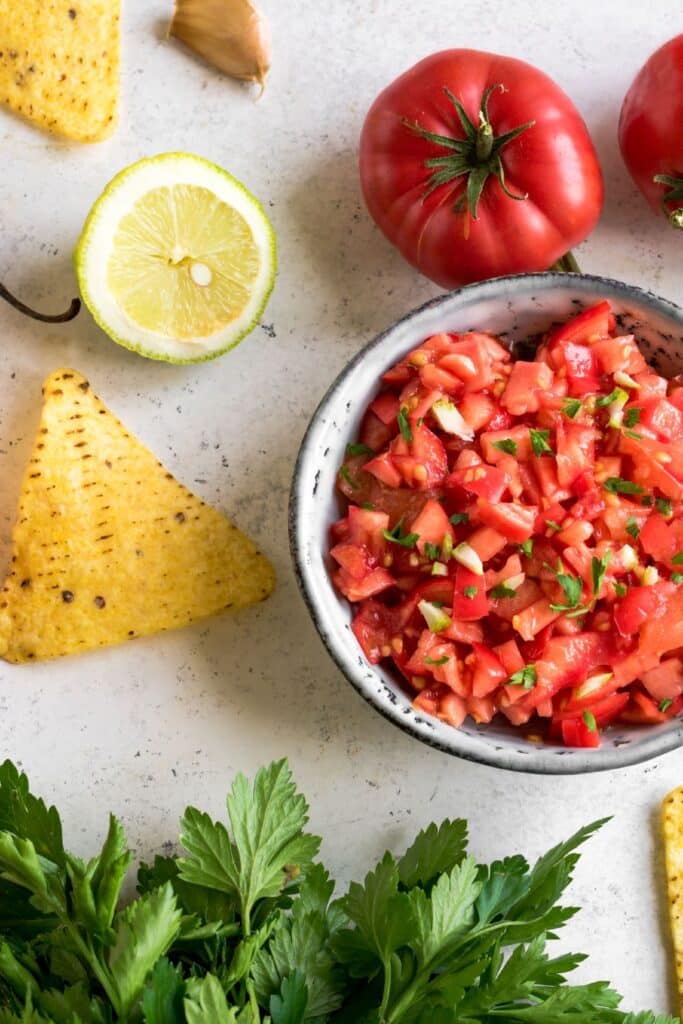 A bowl of Pico de Gallo Fresh Salsa surrounded by tortilla chips, cilantro, lime, and tomatoes.