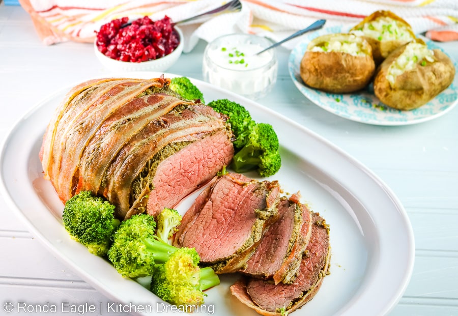 A bacon wrapped eye of round roast on a serving platter with broccoli. The roast has several slices revealing the perfectly medium rare interior