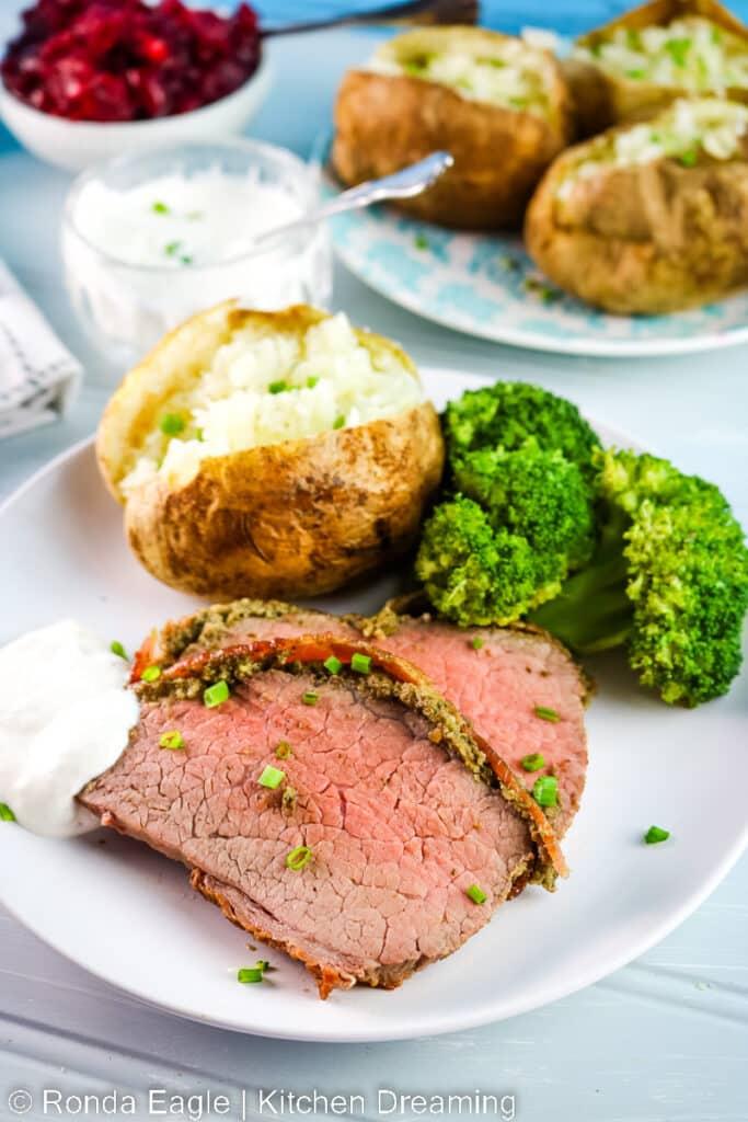 A white dinner plate with a baked potato, steamed broccoli, and slices of bacon wrapped eye of round. A dollop of prepared horseradish sauce sits next to the meat.
