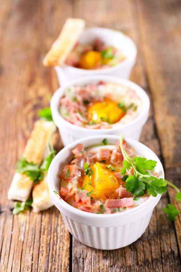 Three white ramekins containing baked eggs with ham and cheese. These are some freshly chopped herbs, whole parsley and toast sticks.