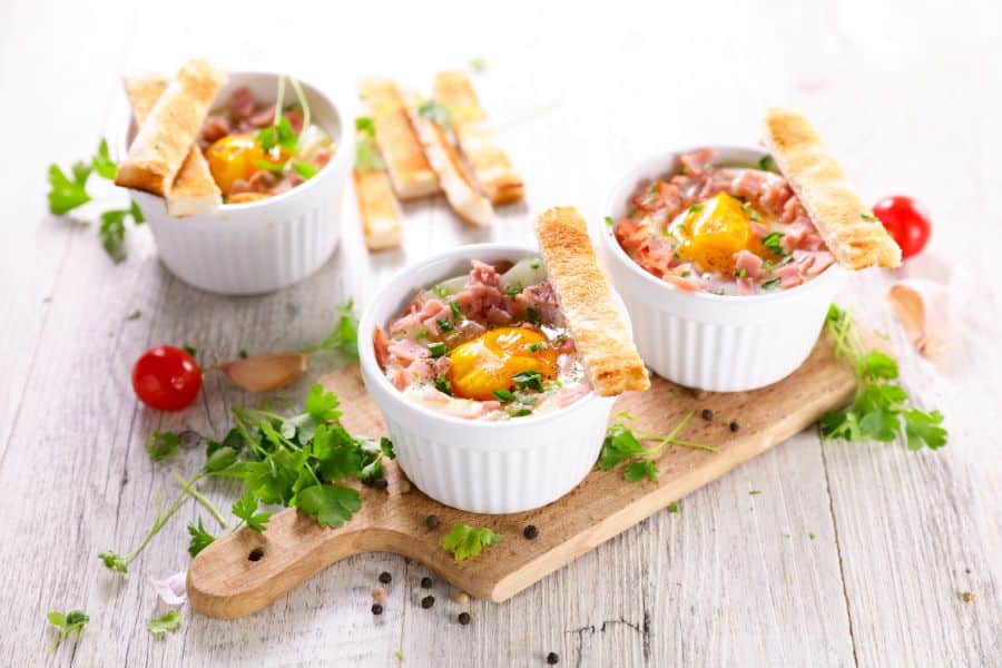 Three white ceramic ramekins containing baked eggs with ham and cheese. These are some freshly chopped herbs, whole parsley and toast sticks.