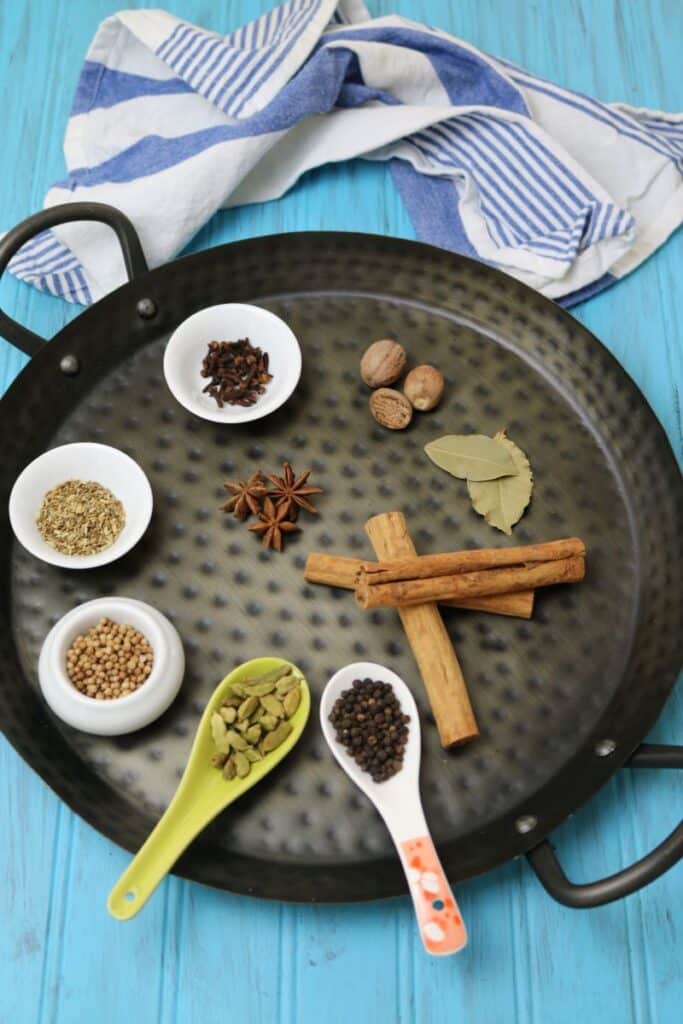 Black hammered metal serving tray with handles holds bowls and loose ingredients for garam masala powder: peppercorns, green cardamom pods. coriander seeds, cumin seeds, whole cloves, bay leaves, cinnamon sticks, nutmeg, and star anise. Some ingredients are optional. 