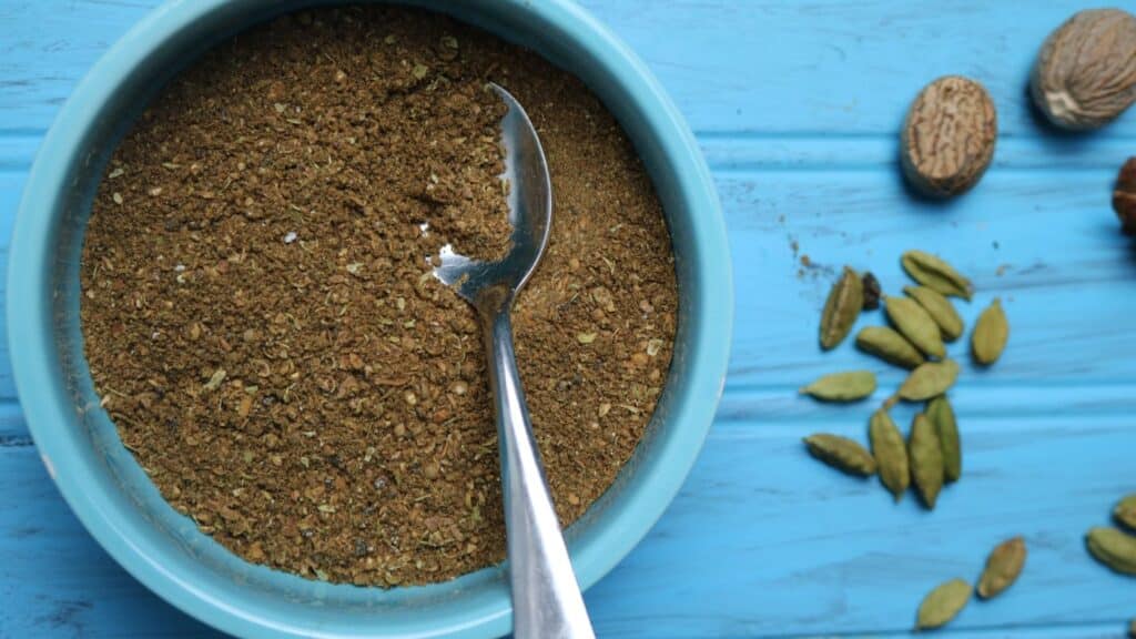 A blue serving bowl filled with freshly prepared Garam Masala Powder. A small silver teaspoon rests inside the bowl. On the blue wooden table top, some whole cardamom pods, star anise, and nutmeg are scattered around the surface. 