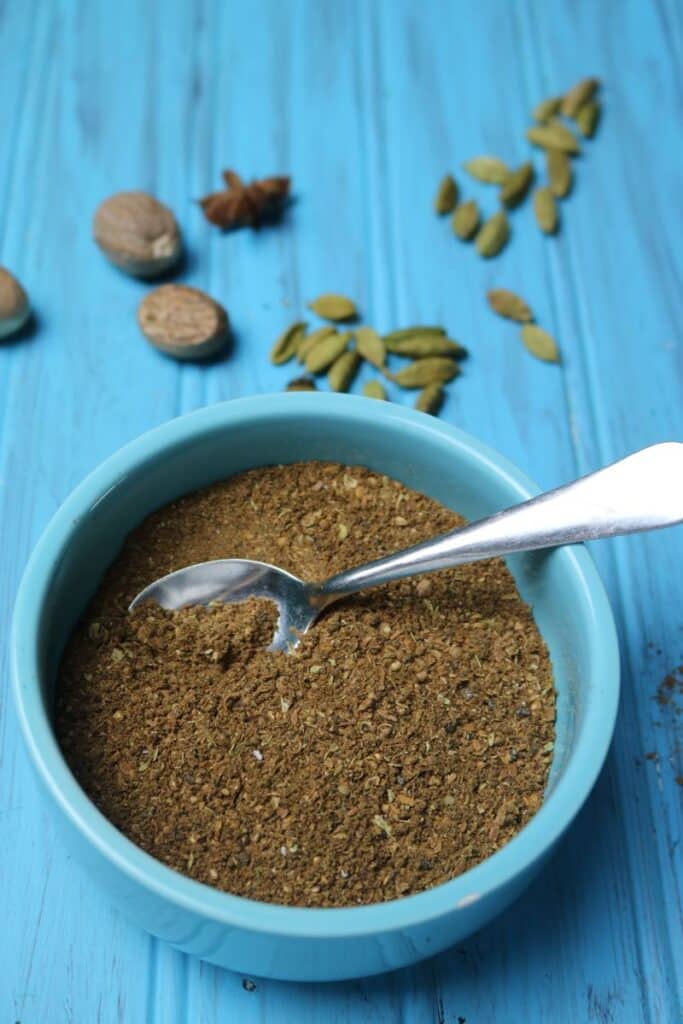 A blue serving bowl filled with freshly prepared Garam Masala Powder. A small silver teaspoon rests inside the bowl. On the blue wooden table top, some whole cardamom pods, star anise, and nutmeg are scattered around the surface. 