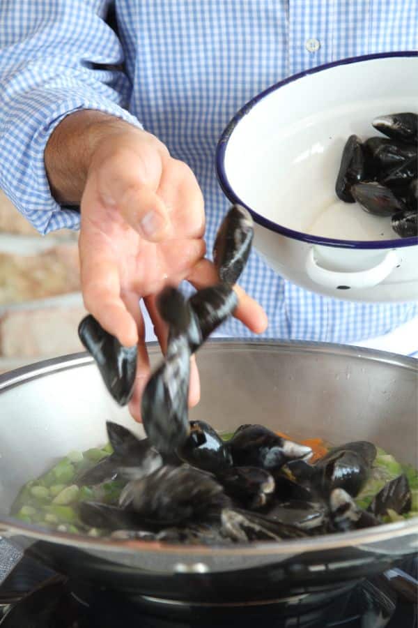 A gentleman prepares mussels in white wine. He is adding mussels into the white wine sauce.