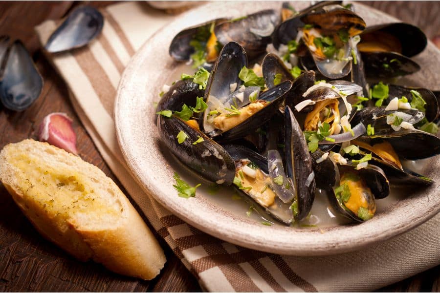 A bowl of mussels in white wine on a table with crusty bread to soak up the delicious white wine broth.