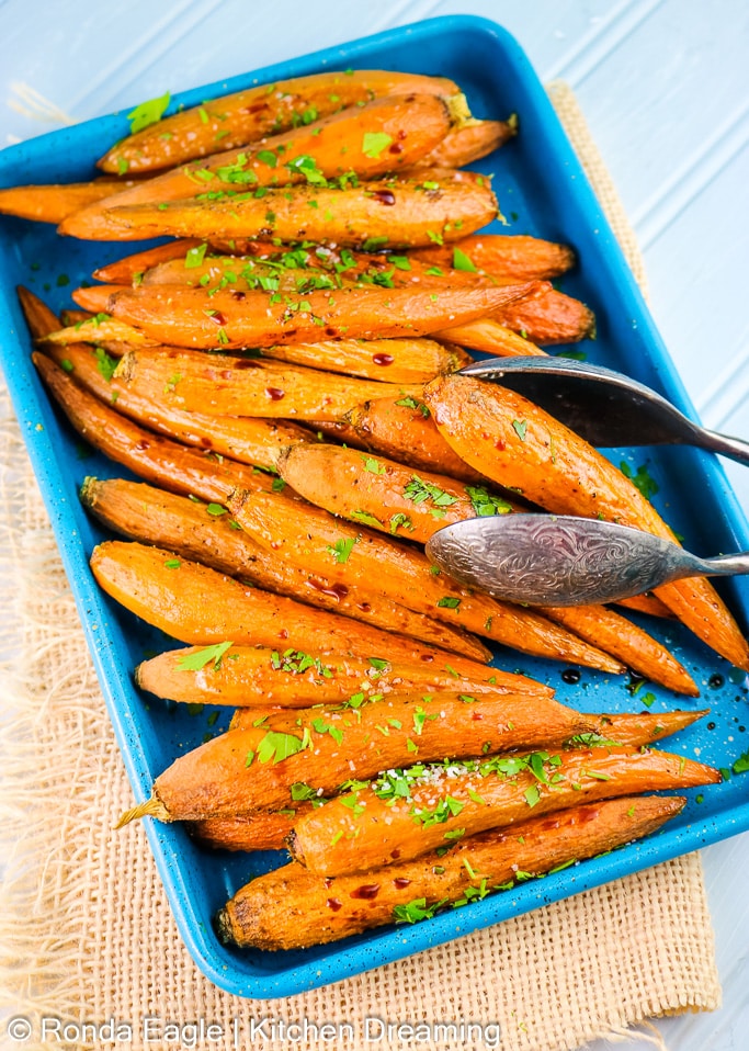 Roasted carrots on a small blue rimmed tray garnished with freshly chopped parsley and a drizzle of balsamic vinegar. A set of vintage metal serving tongs reached into the photo on the right side of the tray.