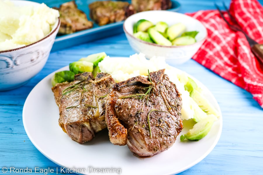 Two roasted lamb chops on a white dinner plate with mashed potatoes and brussel sprouts.