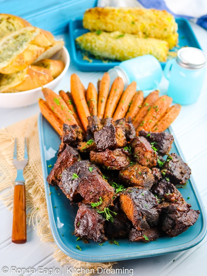 Smoked chuck roast is cubed and coated in tangy BBQ sauce. It is on a blue plate with roasted carrots. In the background, garlic bread and fried corn on the cob is in serving dishes. 