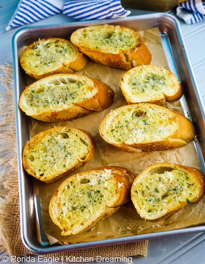 A tray of baked fresh garlic bread slices. You can see the herbs and parmesan cheese.