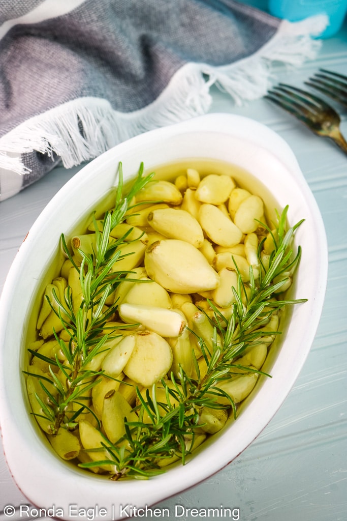 A white oven-safe casserole dish filled with garlic, olive oil, and fresh rosemary sprigs.