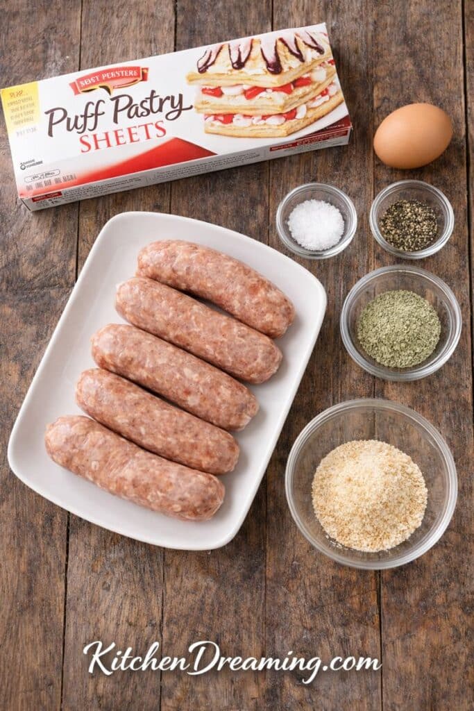 Ingredients for sausage rolls with puff pastry, including whole sausage links, puff pastry, seasonings, and egg wash.