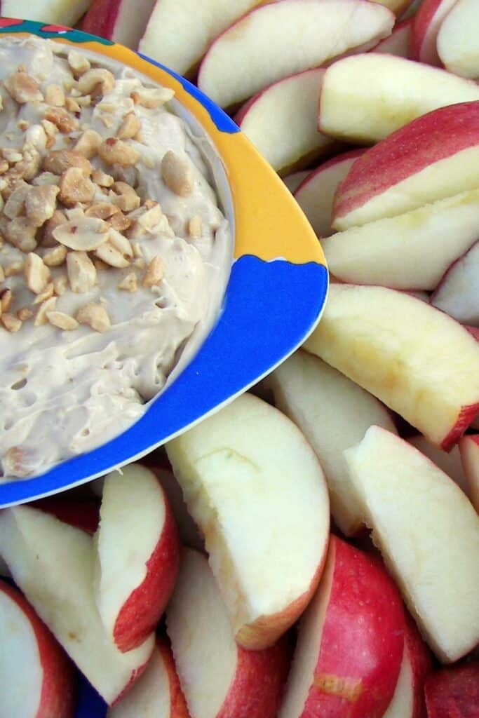 A bowl of cream cheese fruit dip topped with crushed peanuts and surrounded by sliced apples.