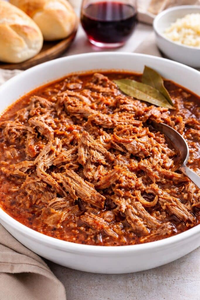 Slow-simmered Portuguese cacoila with tender shredded pork in a rich tomato and red wine sauce, served in a bowl with crusty rolls and rice.