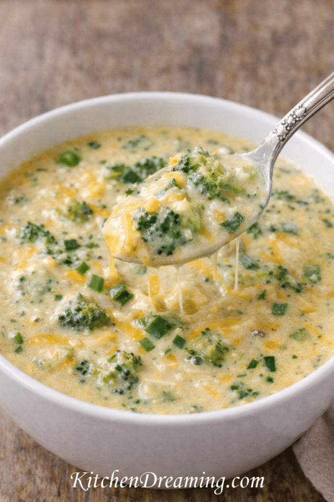 Spoon lifting creamy broccoli cheddar soup with melted cheese and broccoli florets, slow cooker broccoli cheese soup close-up texture.