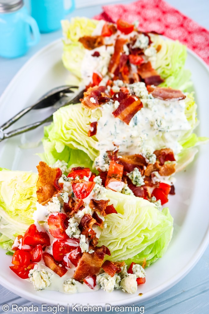 A serving platter of four Iceberg wedge salad topped with creamy herbed ranch dressing, bacon, and cherry tomatoes. 