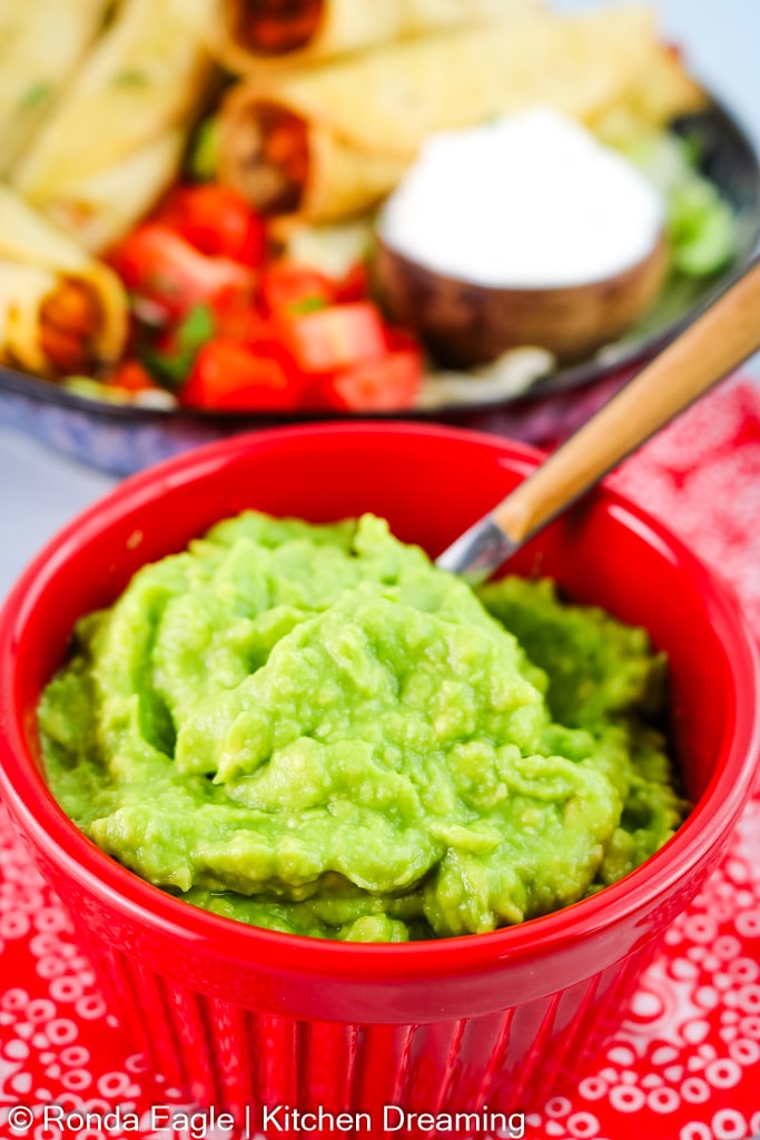 A red bowl filled with vibrant green guacamole is in the foreground. A plate of taquitos is in the background.