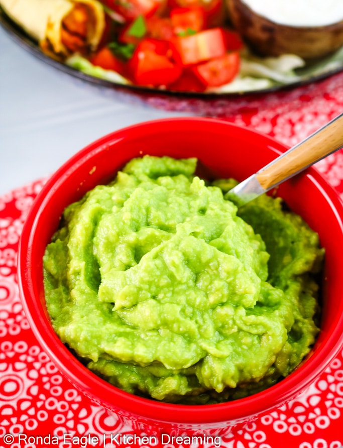 A red bowl filled with vibrant green guacamole is in the foreground. A plate of taquitos is in the background.