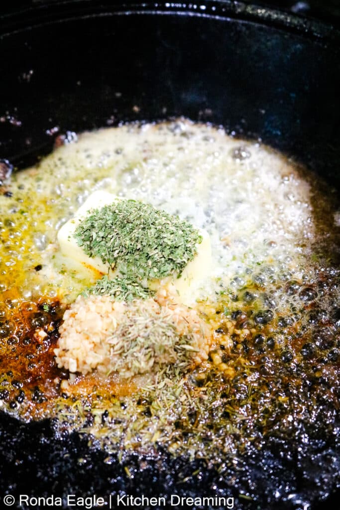 A cast iron skillet containing butter, herbs, and spices to create the sauce for the butter garlic steak bites.