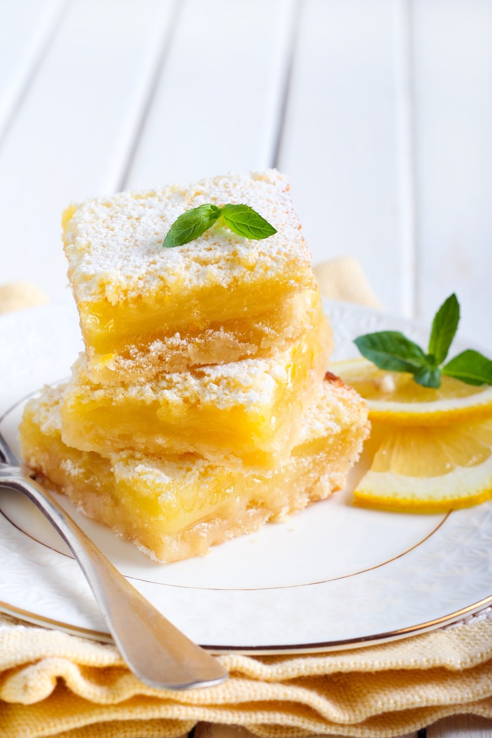 A stack of 3 lemon bars on a plate with a fork and some lemon slices and mint garnish.