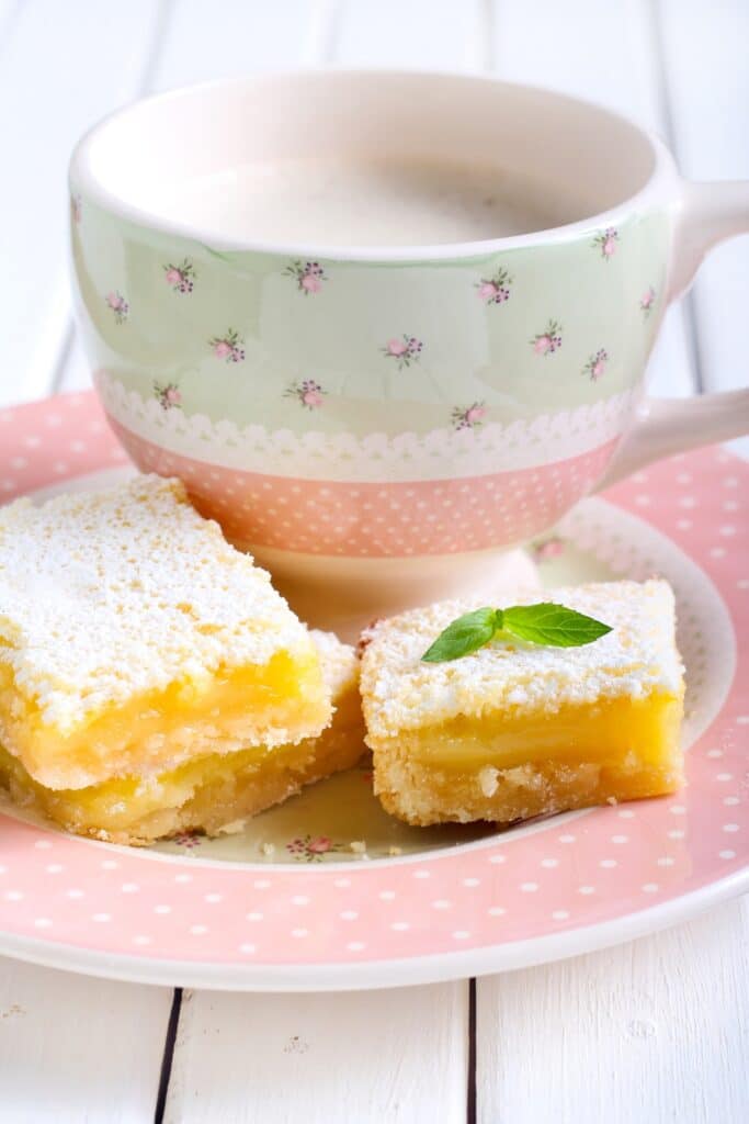 Three small lemon bars sitting on a small tea saucer. A cup of tea in the background.