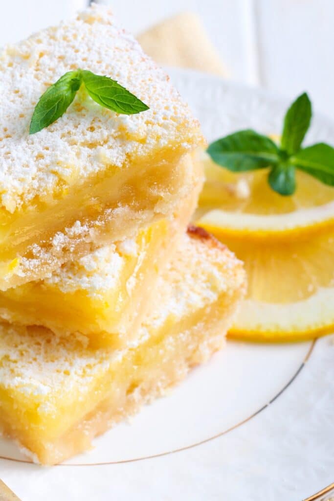 A close up image of three lemon bars arranged in a stack on a white plate. The lemon bars are dusted with powdered sugar and garnished with mint.