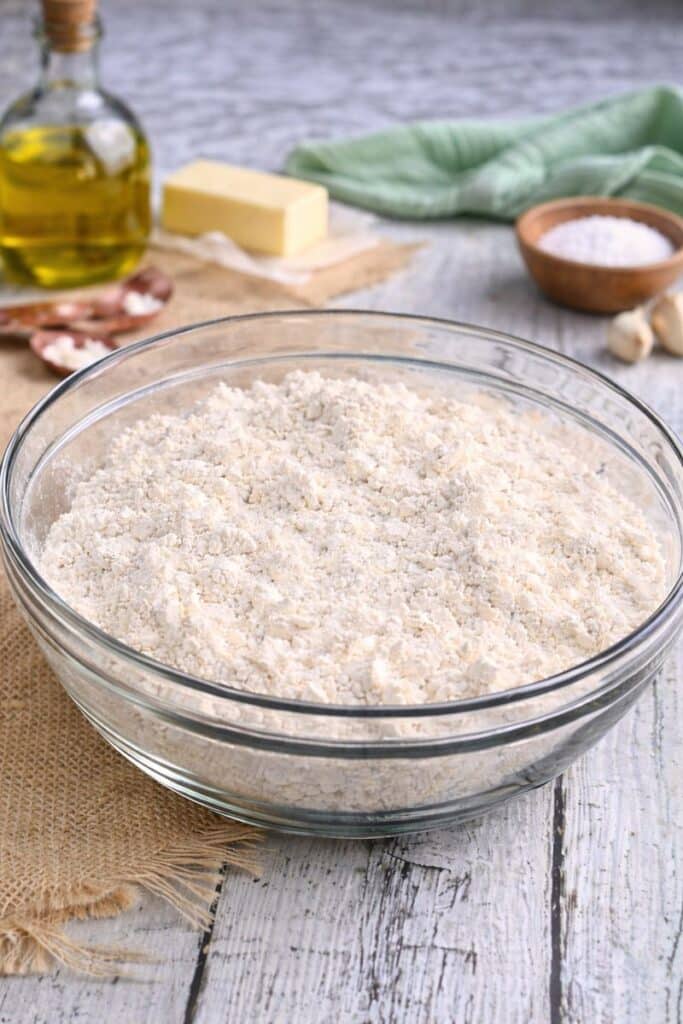 Homemade Baking Mix (Easy Bisquick Substitute with 5 Pantry Ingredients) 2 Homemade baking mix in a clear glass bowl on a rustic kitchen surface, with butter, oil, and pantry ingredients in the background.