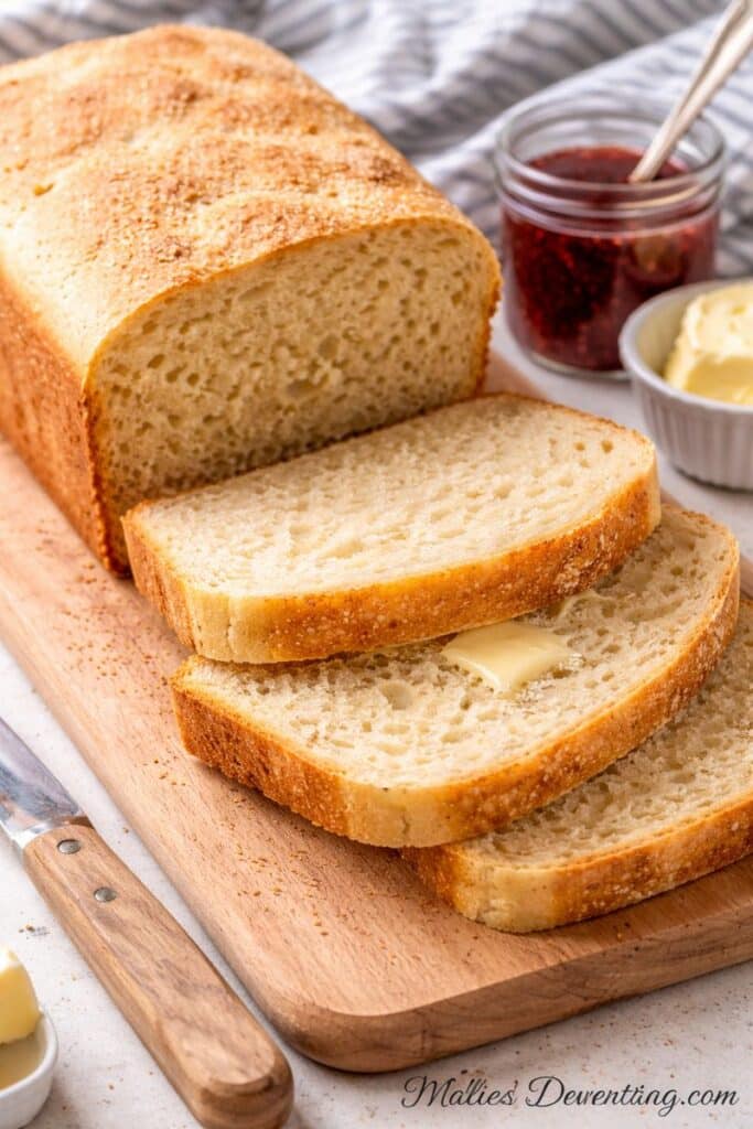 Bread machine English muffin bread sliced and stacked on a cutting board with butter and jam, showing a soft nooks-and-crannies texture.