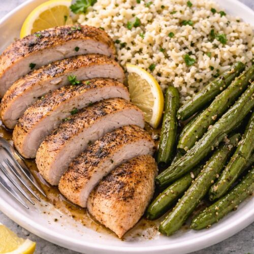 Juicy oven-baked chicken breast sliced and served with rice, green beans, and lemon on a plate, showing a healthy baked chicken dinner.