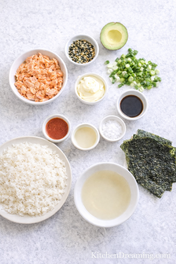 Salmon Sushi Bake Recipe (Easy, Creamy Sushi Bake) 3 Ingredient flat lay for salmon sushi bake recipe with bowls of sushi rice, cooked salmon, mayonnaise, furikake, sauces, and nori.